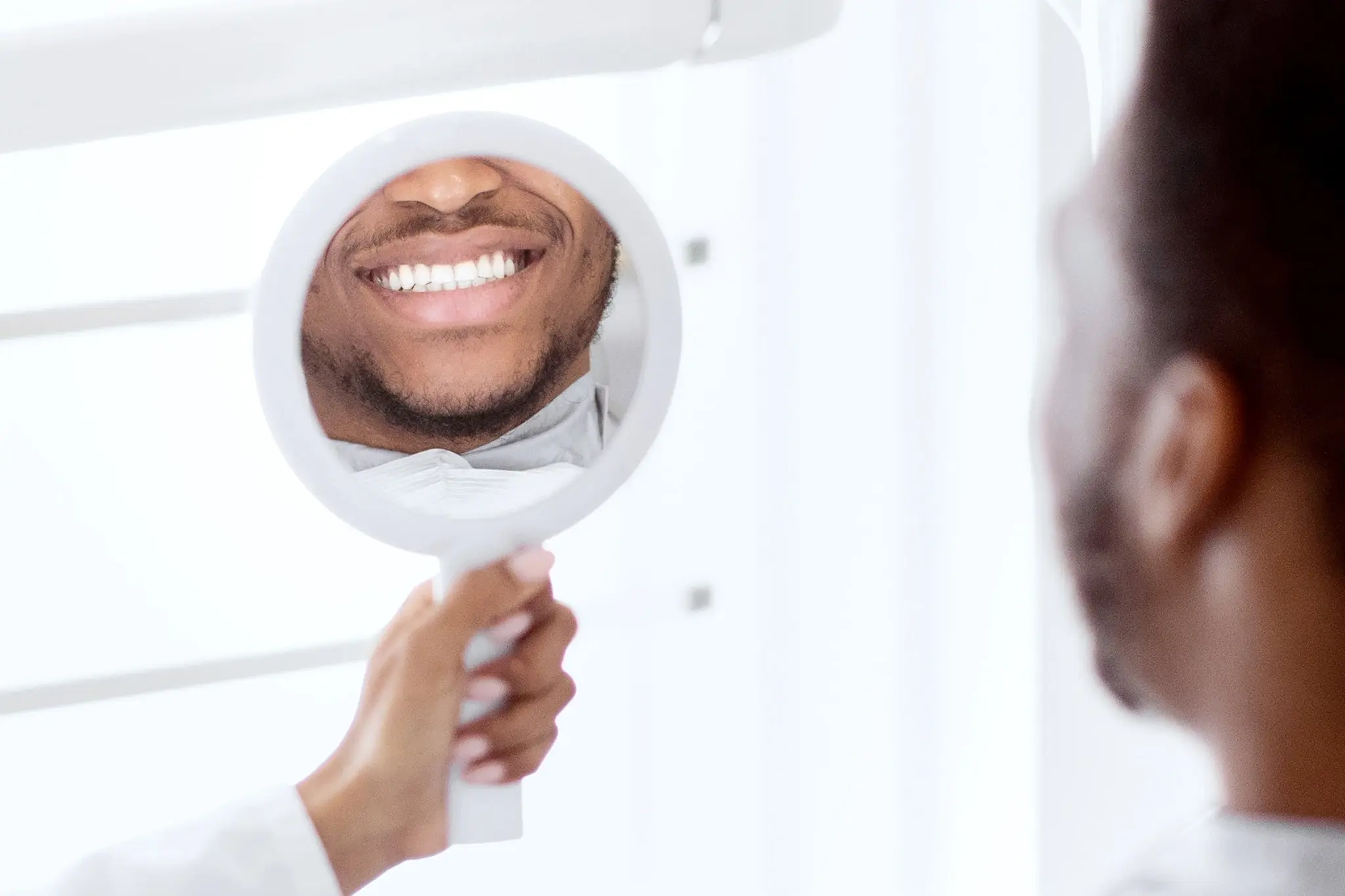 A black man looks through a mirror held up by his dentist, checking his teeth with a smile.