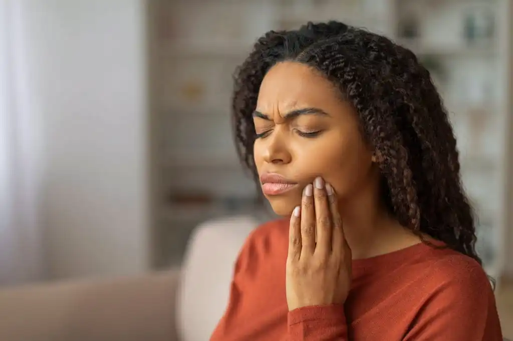 A woman with curly dark hair, wearing a dark orange top, sits indoors with eyes closed, holding her cheek with her hand. She has an expression of pain and appears to be experiencing a toothache.